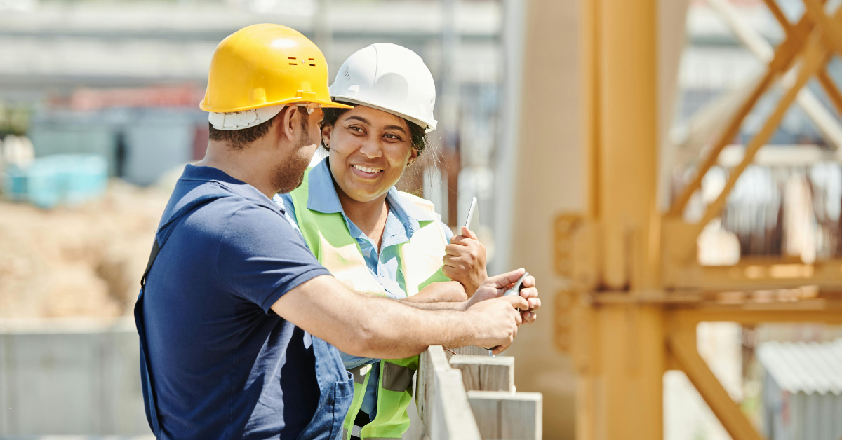 Two construction workers wearing hard hats and safety vests, standing on a construction site. They are engaged in conversation.